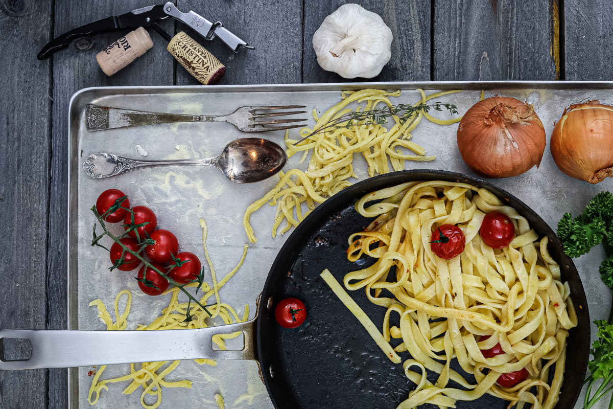Pasta aus Hartweizengrieß_1 Pfanne mit Nudeln und Kirschtomaten auf Blech, daneben Knoblauch, Zwiebeln, Tomatenzweig, Löffel, Gabel und Weinöffner.