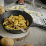 Bratkartoffeln in einer gusseisernen Pfanne mit Gabel auf Holzbrett, umgeben von rohen Kartoffeln und Küchentuch.