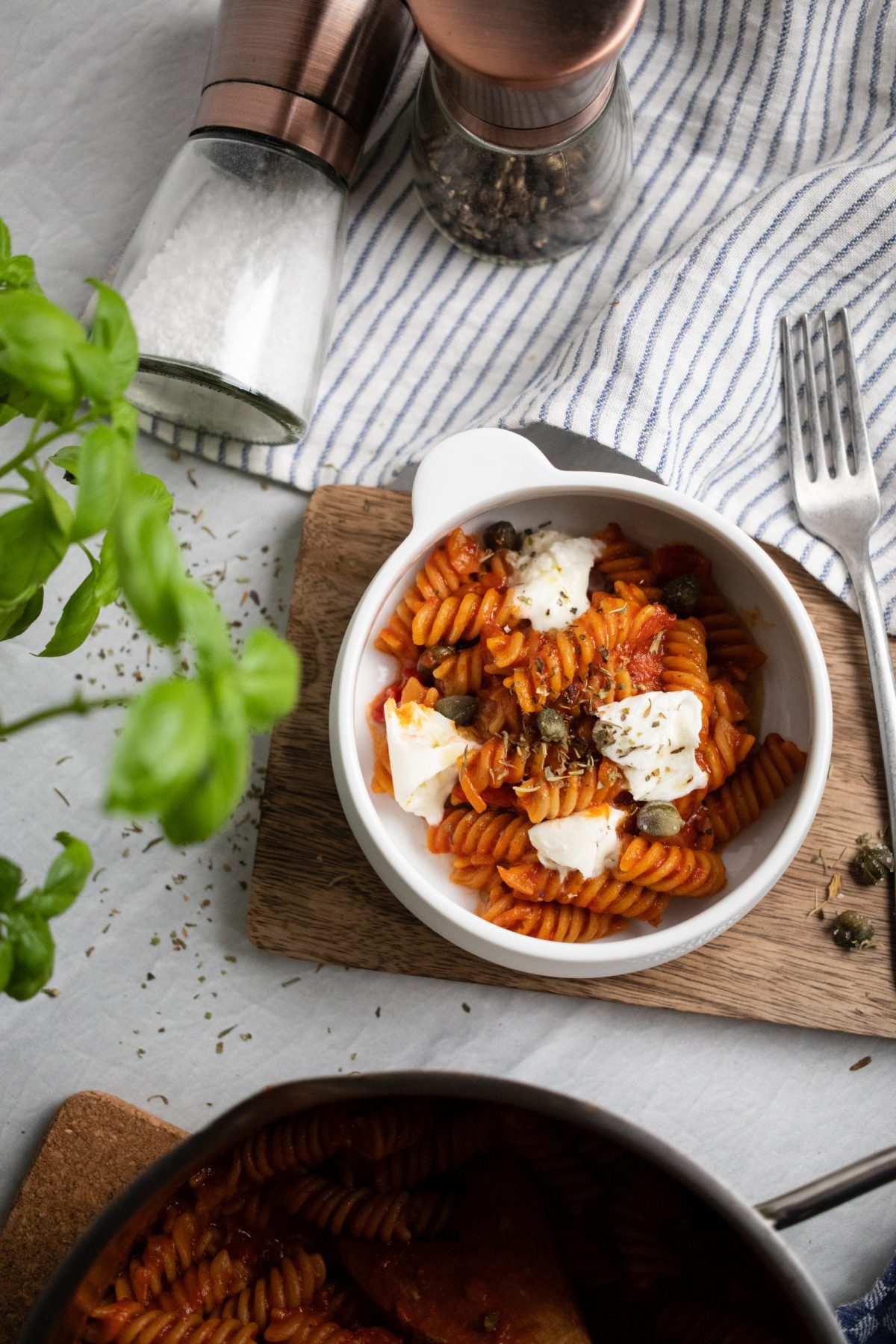 Schüssel mit Fusilli in Tomatensauce, garniert mit Käse und Kapern, neben Basilikum und Gewürzmühlen auf gestreiftem Tuch.