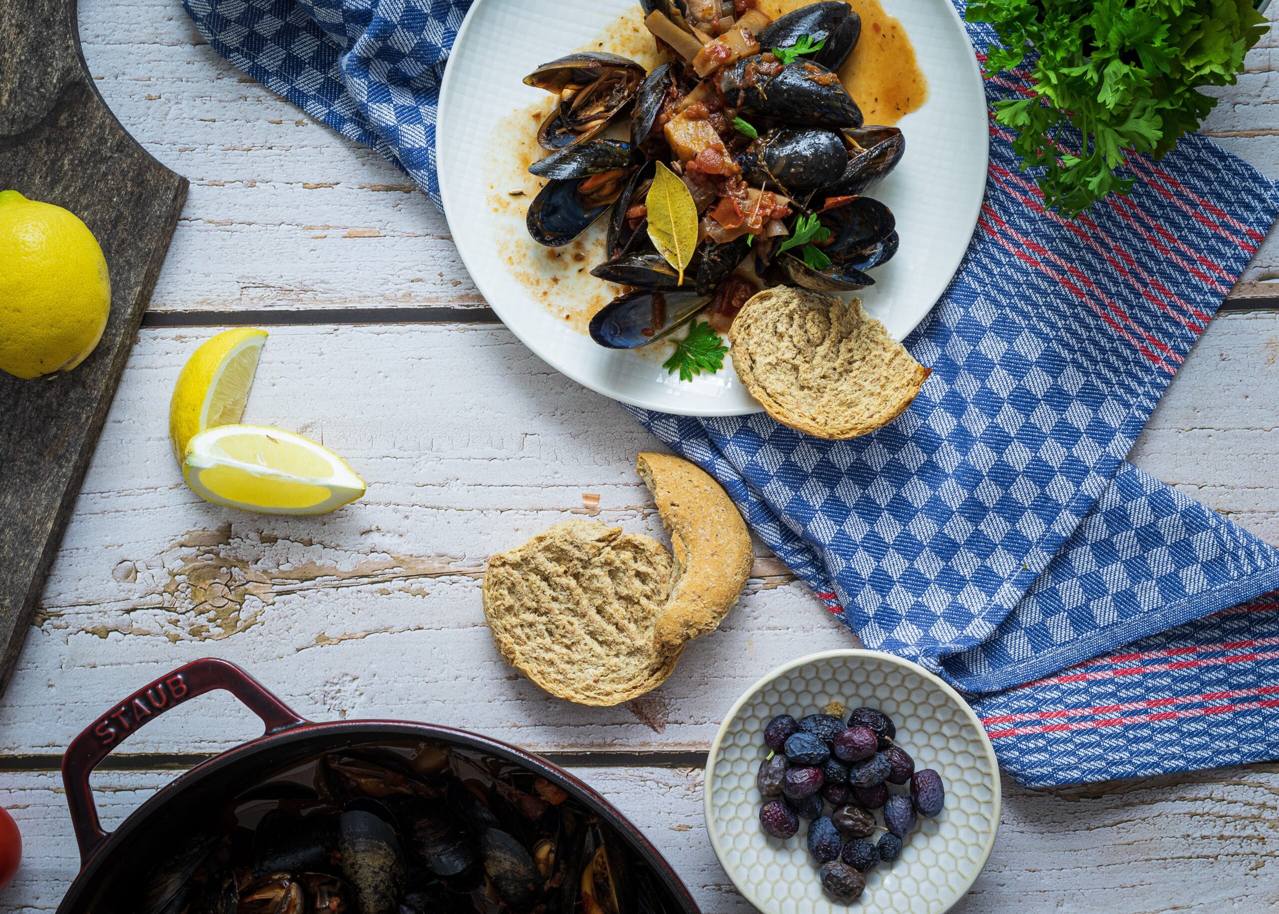 Teller mit Muscheln und Brot auf blauem Geschirrtuch, Zitronenscheiben und Olivenschale daneben auf Holztisch.