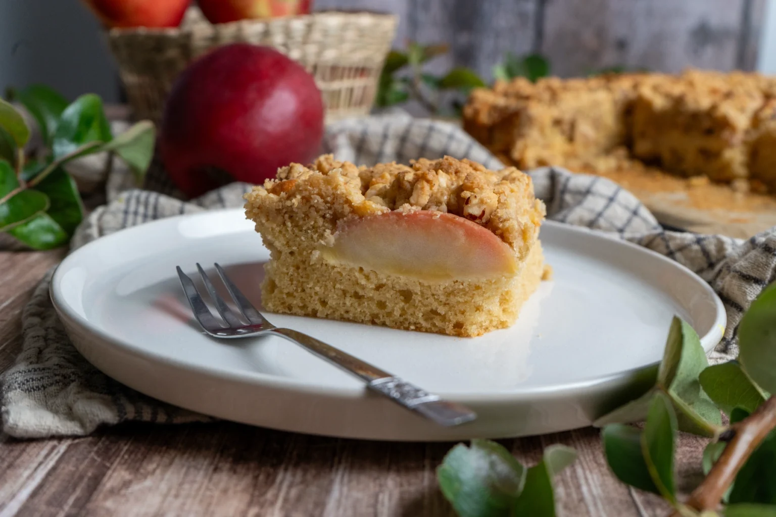 Stück Apfelkuchen mit Streuseln auf einem weißen Teller, Gabel daneben, im Hintergrund ein Korb mit Äpfeln.