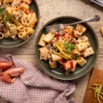 Zwei Teller Rigatoni mit Hackfleischsoße, garniert mit Kräutern und Gurke. Daneben Schalotten und Brett mit Zutaten.