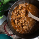 Gusseisener Topf mit Rindergulasch, Holzlöffel, grüne Blätter und graues Tuch daneben auf Holztisch.