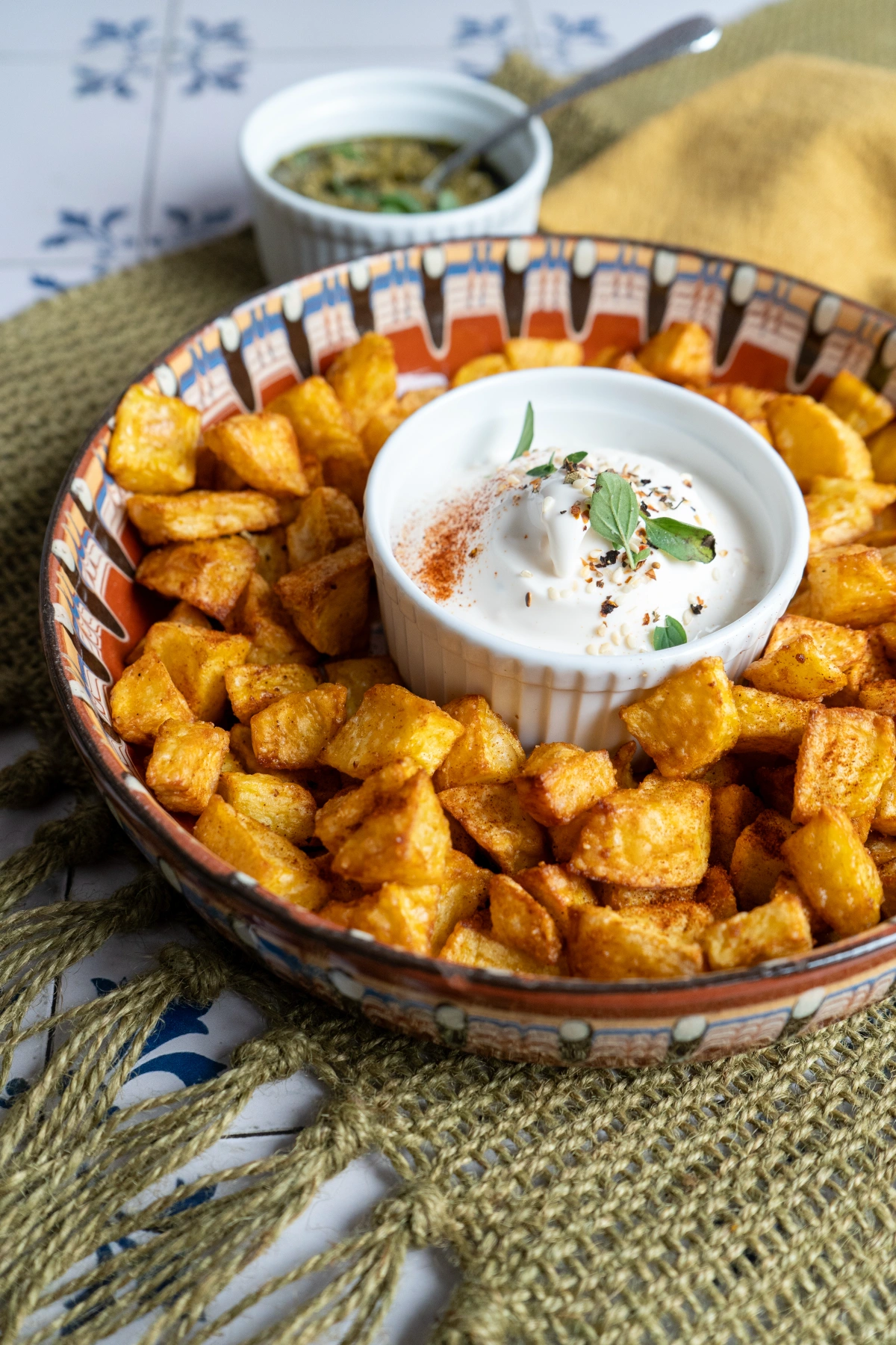 Knusprige Kartoffelwürfel aus der Heißluftfritteuse-04 Knusprige Kartoffelwürfel in Schale mit Dip in der Mitte, dekoriert mit Kräutern, auf grünem Tuch serviert.
