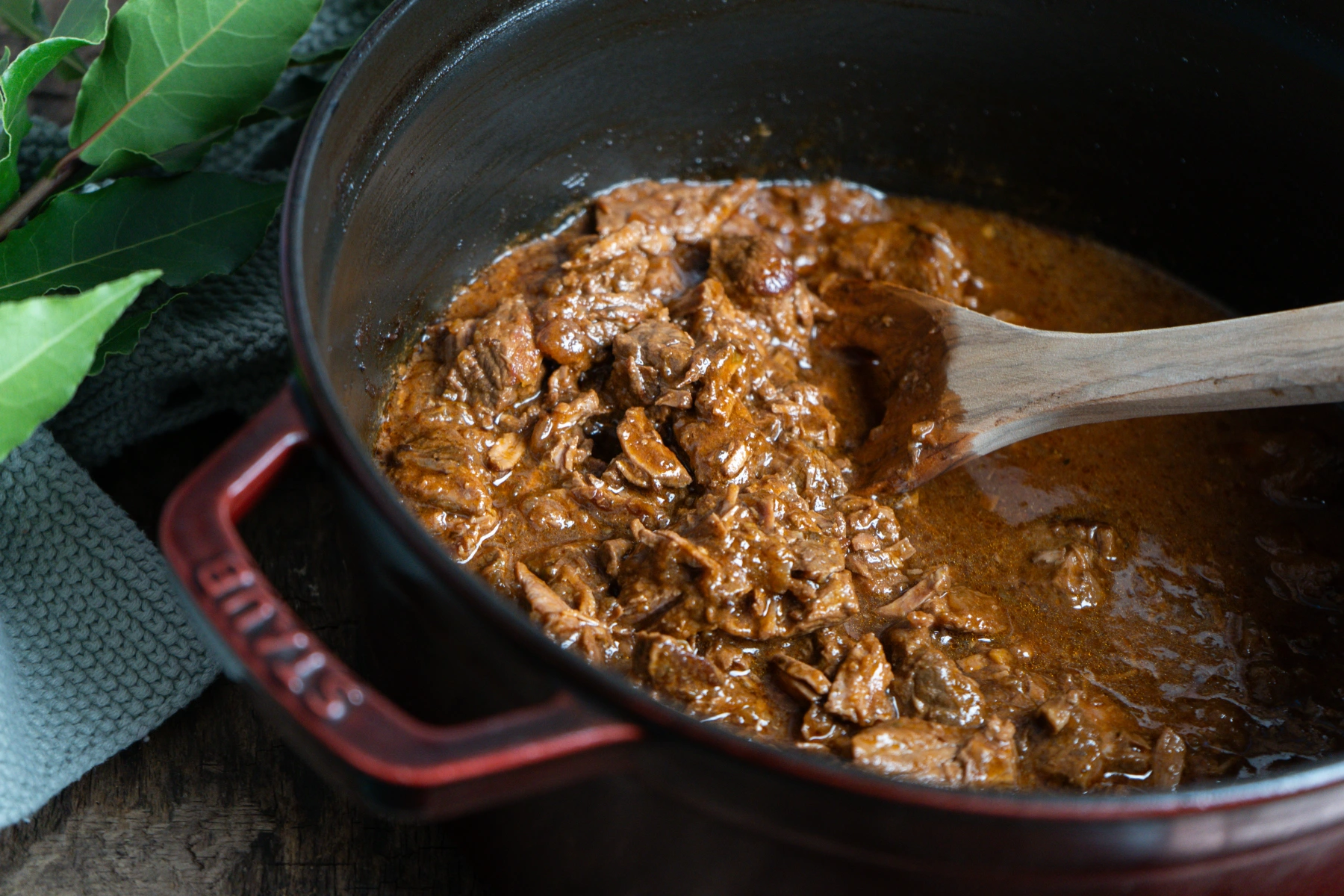 Wiener Saftgulasch-02 Rotes Gusseisen-Topf mit Rindfleisch-Gulasch, angerührt mit Holzlöffel, daneben Lorbeerblätter.