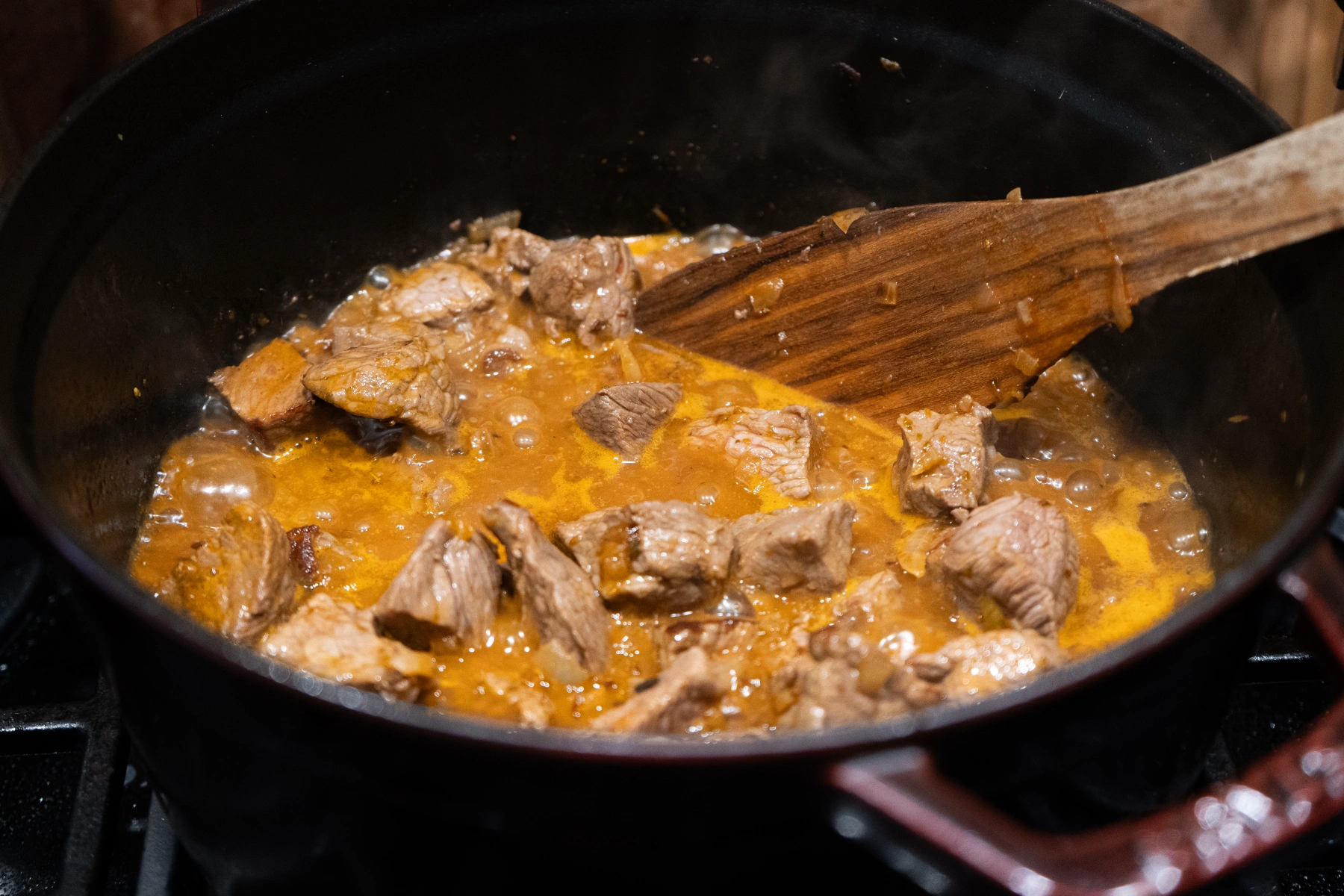 Wiener Saftgulasch-06 Fleischstücke in brauner Soße blubbern in einem Topf. Ein Holzlöffel rührt die Mischung um.