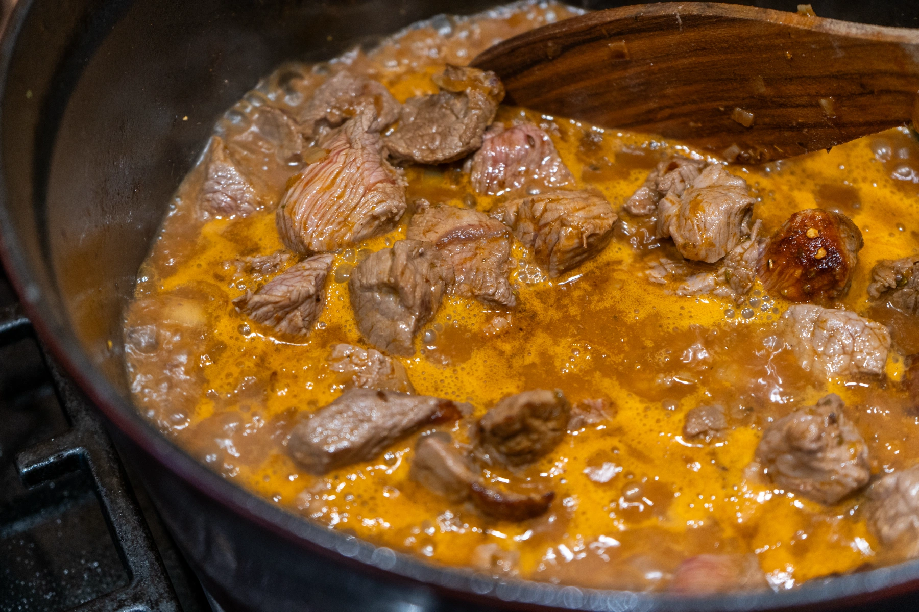 Wiener Saftgulasch-07 Stückiges Rindfleisch in einer köchelnden Tomatensoße, angerührt mit einem Holzlöffel in einem Topf.