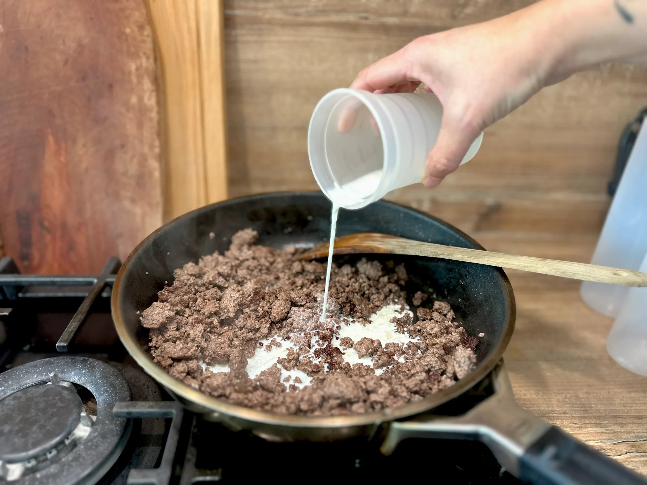 Hand gießt Flüssigkeit aus einem Becher in eine Pfanne mit gebratenem Hackfleisch auf einem Herd.