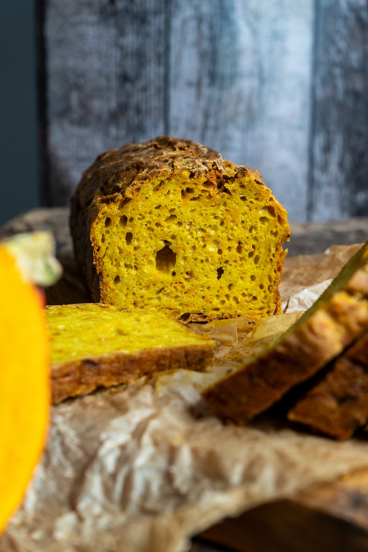 Würziges Kürbisbrot-02 Nahaufnahme eines geschnittenen Kürbisbrots auf Backpapier, im Hintergrund eine rustikale Holzoberfläche.